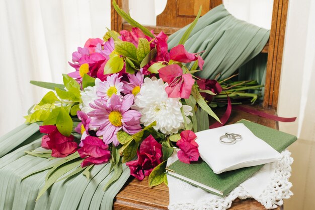 Обручальные кольца на подушке над книгой возле букета цветов и свадебное платье на стуле
