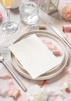 Место свадебного стола с пустой карточкой около цветов, шелковых лент и пер, крупного плана. романтическая сцена с вертикальным макетом бумажной карты на тарелке на мраморном столе