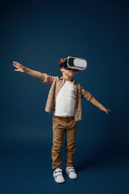 Weightless in the sky. Little boy or child in jeans and shirt with virtual reality headset glasses isolated on blue studio background. Concept of cutting edge technology, video games, innovation.