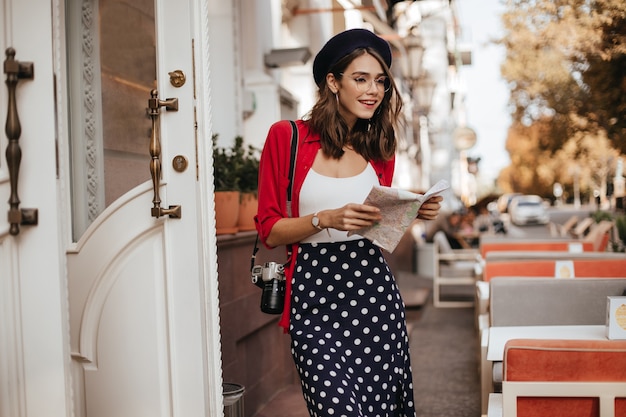 Free photo well-built young lady with brunette hair, long polka dot skirt, white blouse, red shirt, beret and glasses walking on town with map in hands and camera during daytime