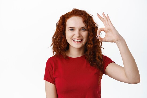 Well done. Smiling confident teen ginger girl showing OK sign, approve and agree, praise excellent choice and looking satisfied, standing in t-shirt against white wall