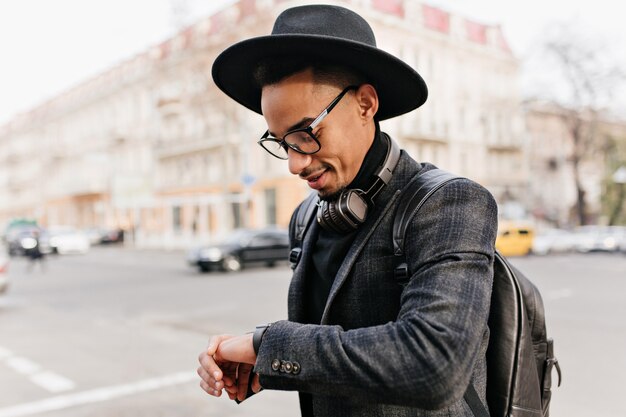 彼の腕時計を見ている身なりのよいアフリカの紳士。帽子をかぶったムラートの男の屋外写真は革のバックパックを身に着けています。