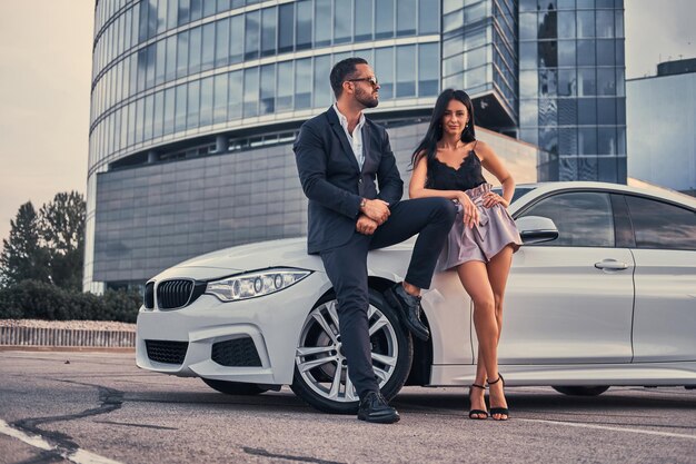 Хорошо одетая привлекательная пара, опирающаяся на роскошный автомобиль на открытом воздухе против небоскреба.