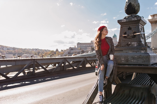 Well-dressed female tourist drinking coffee on city background in windy day