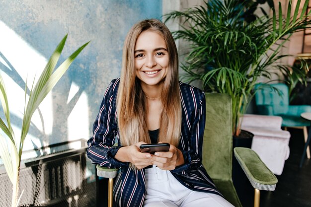 Well-dressed interested woman sitting in front of big plant. Appealing laughing girl with phone chilling in arm-chair.