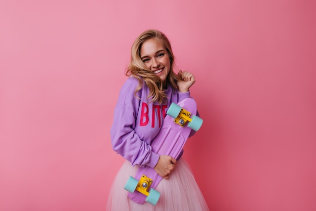 Well-dressed lady with skateboard smiling on pink backgorund. Inspired caucasian girl with blonde hair holding longboard.