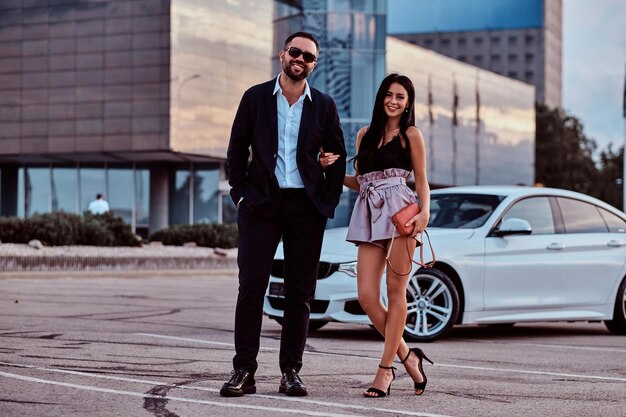 Хорошо одетая улыбающаяся пара вместе позирует возле роскошного автомобиля на фоне городского пейзажа.