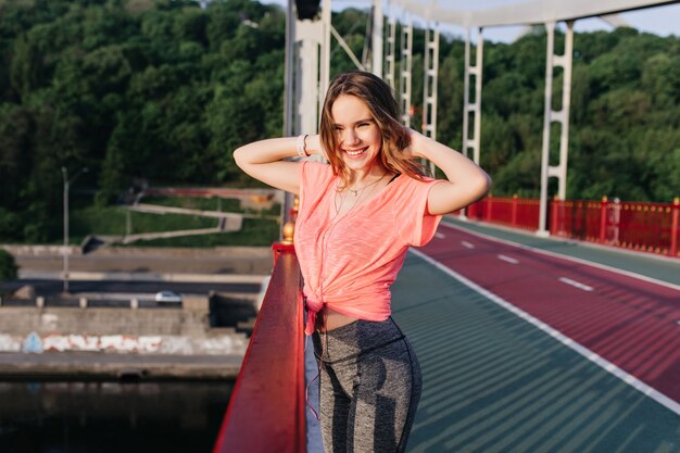 Well-dressed young lady posing at cinder path with inspired smile. Excited blonde girl standing with hands up at stadium.