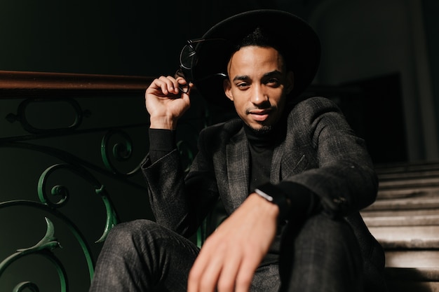 Well-dressed young male model posing on steps with gently smile. Indoor photo of curious african guy wears hat.