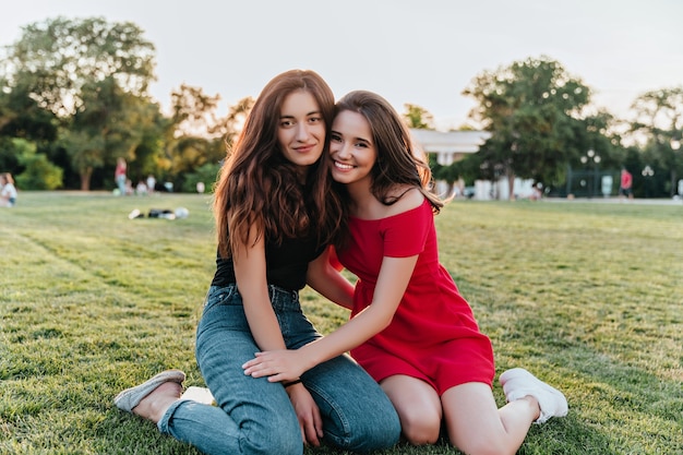 Well-dressed young sisters sitting on the grass in warm spring day. Female best friends posing together in beautiful park.