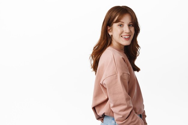 Wellbeing and people concept. Portrait of young smiling redhead woman turn head at camera, stands in profile, looking happy and friendly, standing against white background.