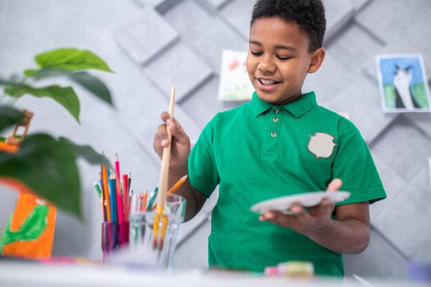 Wet brush. Joyful cute dark-skinned boy of school age with palette lowering brush into glass of water standing near table with colored pencils in bright room