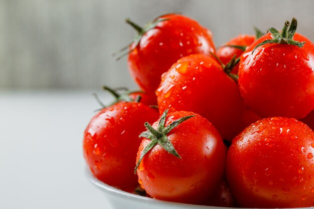 Wet juicy tomatoes in a white plate on a white and grungy surface. side view.