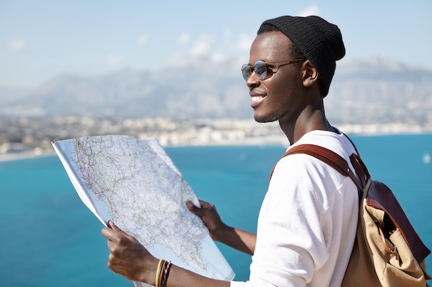 What a beautiful landscape! Happy excited Afro American backpacker using paper map while standing on viewing point high above blue sea and studying surroundings during his trip. Travel and adventure