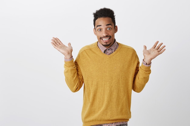 What done cannot be changed. Portrait of confused unaware emotive dark-skinned guy with afro hairstyle raising palms in surrender, shrugging and smiling cluelessly, having no idea over gray wall