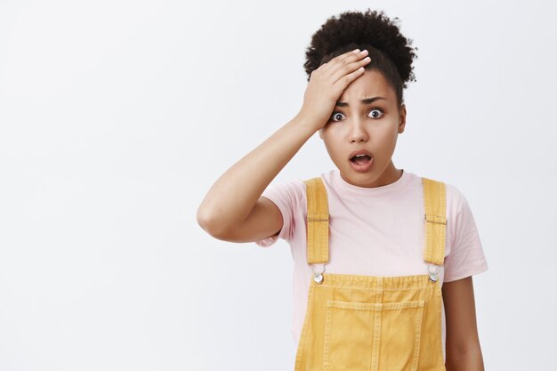 What have you done. Portrait of shook displeased and upset African American sister in yellow overalls, holding palm on forehead, dropping jaw and frowning, being disappointed and distressed