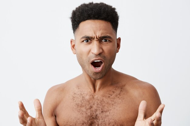 What the heck. Close up of beautiful black-skinned man with curly hair without clothes spreading hands with confused expression, when his favourite football team losing match