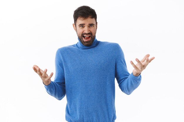 What the hell happening. Portrait of confused and frustrated, bothered caucasian guy, raise hands spread in dismay, staring disappointed and upset, complaining at bad thing, white wall