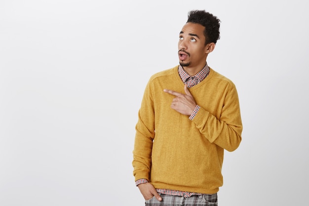 What the hell is that. Indoor shot of questioned curious african male with afro haircut pointing and looking at upper left corner, seeing interesting thing and discussing it with friends