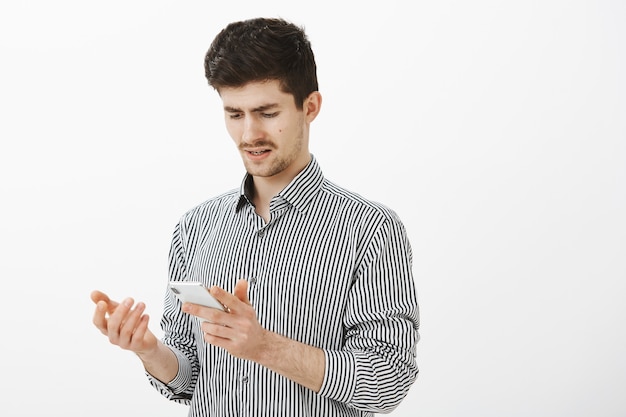 What the hell is that. Portrait of confused frustrated attractive male student with moustache in striped shirt, holding smartphone and gesturing with palm, being doubtful and frustrated over gray wall