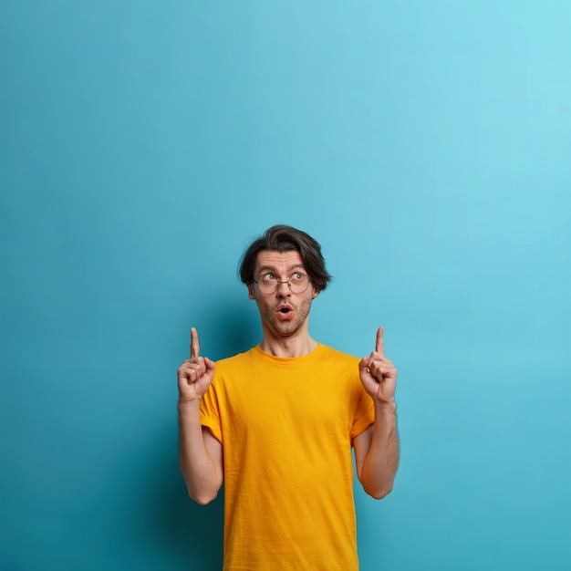 What incredible copy space. Wondered hipster keeps mouth opened, points both index fingers upwards, surprised and interested in great deal, wears yellow t shirt, gestures against blue wall