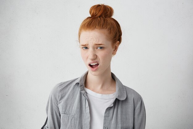 What? Indignant young beautiful woman with hair knot dressed in casual clothing standing at grey wall, grimacing, expression her dislike or disregard, feeling unwillingness towards something