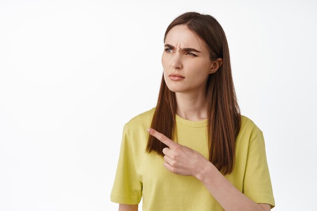 What is that. Confused brunette girl, pointing and looking left with frowning suspicious face, see something strange, questioned by promo, white background.