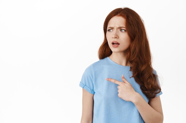 What is that. Confused young redhead woman frowning puzzled, pointing and looking left at something strange, stare puzzled at copy space for your logo or advertisement, white background.