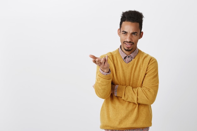 What is your point, dude. Portrait of displeased dark-skinned male office worker in yellow pullover holding hands half-crossed, pointing aside with palm, expressing contempt and dislike, arguing