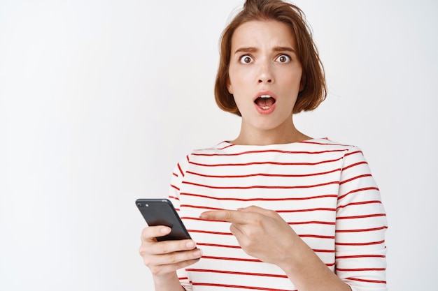 What it that. Shocked and worried woman pointing at smartphone and look anxious , stare confused, cant understand something on mobile phone, white wall