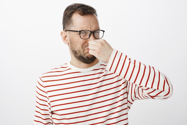 Free photo what nasty smell. portrait of displeased disgusted funny guy in glasses, covering nose with fingers and frowning from displeasure, smelling awful reek