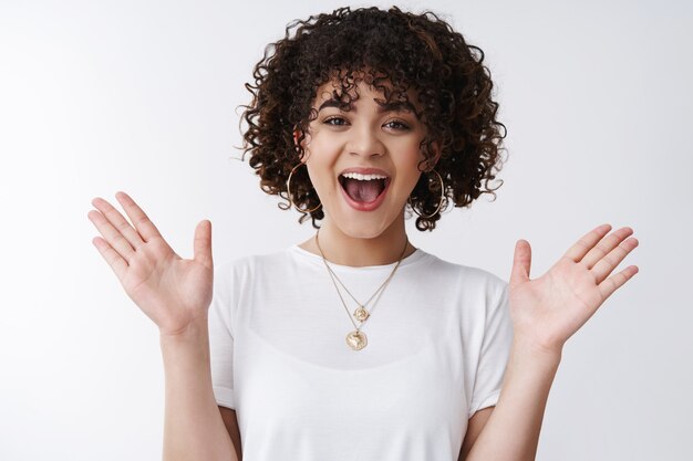 What nice surprise. Amazed happy friendly charming curly-haired brunette female inviting coworker glad see friend smiling raising hands amazed standing amused happy entertained, white background