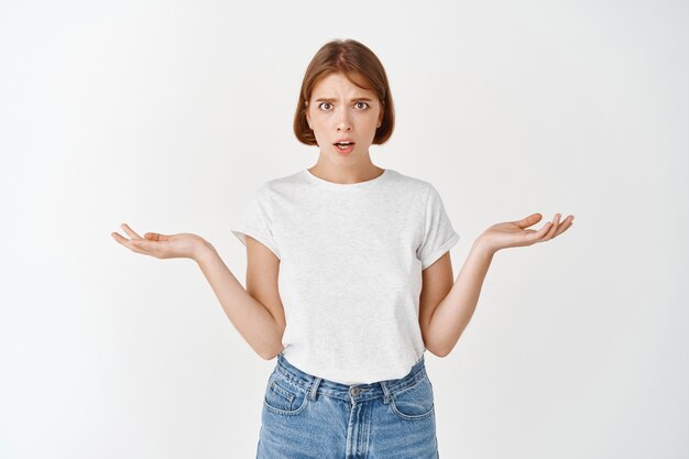 Che problema. giovane donna confusa con i capelli corti, alzando le spalle con le mani vuote di lato, guardando preoccupata e perplessa, muro bianco,