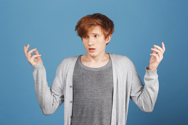 What's going on. Young handsome redhead male student in casual grey outfit spreading hands with lost expression, don't understanding what going on on exam