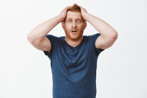 What should I do now. Portrait of shocked and desperate attractive redhead with bristle, holding hands on hair, popping eyes and dropping jaw from shock and despair, standing over gray wall