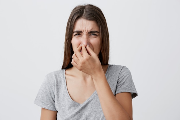 What a smell. Close up of young unhappy caucasian student girl with long hair in casual t-shirt squeezing nose with fingers,  with disgust expression