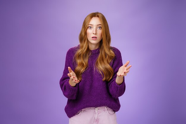 What wrong, I worried. Portrait of nervous and empathical young redhead woman feeling anxious raising hands questioned and looking at camera wondered expressing concern for friend over purple wall.