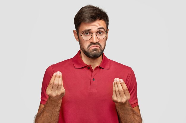 What do you want from me? Bearded young male with displeased expression, shows Italian gesture wtf, wears spectacles and red t-shirt, stands over white wall. Man shows interrogative gesture