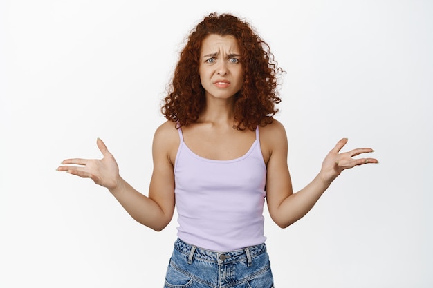 Free photo whats happening. frustrated redhead girl, shaking hands and shrugging confused, cant understand, staring puzzled and upset on white.