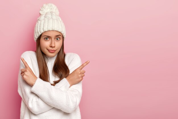 Which one to choose. Hesitant natural European woman points sideways, crosses arms over chest, suggests two variants, makes decision, wears warm white sweater and hat, blank space on pink background