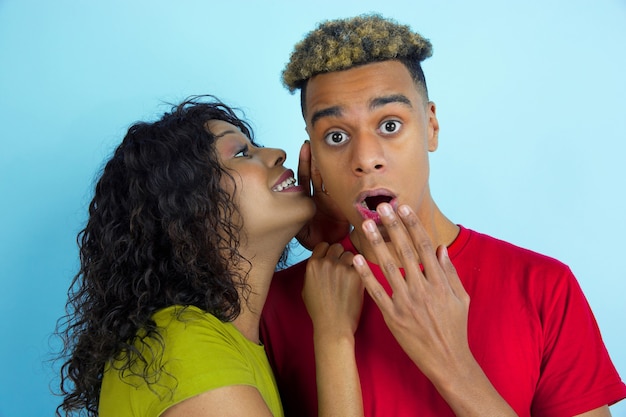 Бесплатное фото Шепчет секрет. молодой эмоциональный афро-американский мужчина и женщина в красочной одежде на синей стене. прекрасная пара.