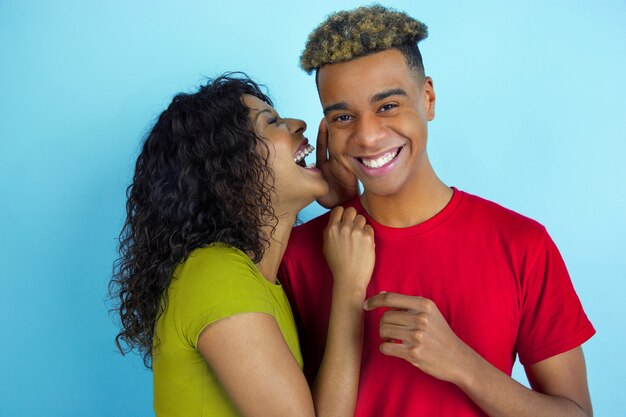 Whispering a secret. Young emotional african-american man and woman in colorful clothes on blue wall. Beautiful couple.