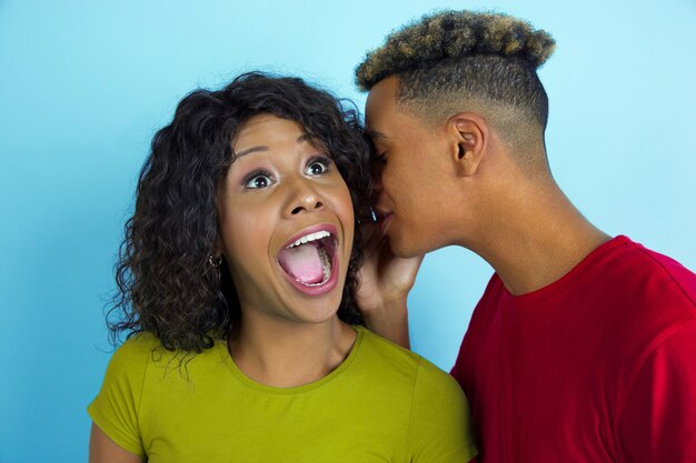 Whispering a secret. Young emotional african-american man and woman in colorful clothes on blue wall. Beautiful couple.