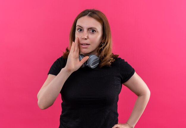 Foto gratuita sussurrando la giovane donna casuale che indossa le cuffie sul collo con la bocca del neard della mano sulla parete rosa isolata