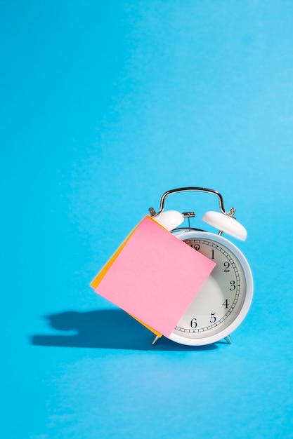 Free photo white alarm clock and blank pink paper sticker on a blue background isolated