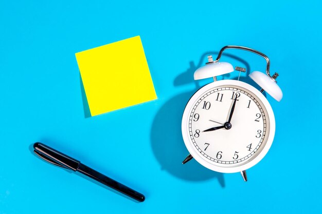 white alarm clock and blank yellow paper sticker on a blue background isolated