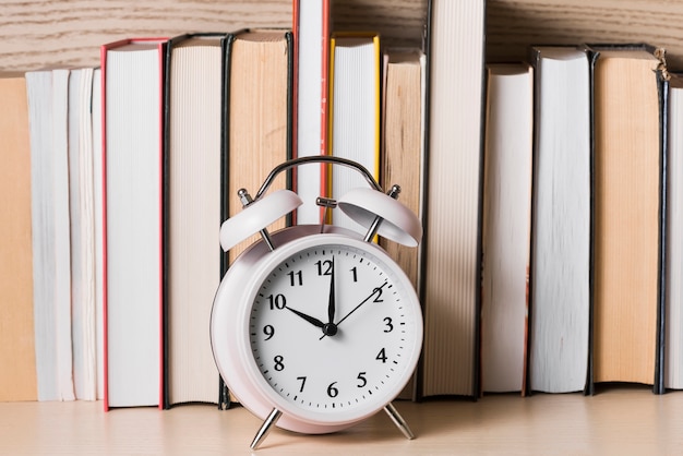 Free photo white alarm clock showing 10'o clock in front of bookshelf on wooden desk