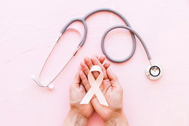 Free photo white awareness ribbon in two hands with stethoscope on pink background