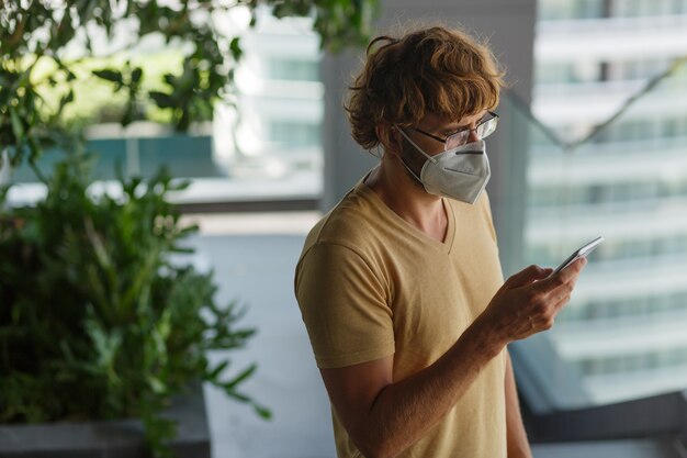 White bearded adult man using smartphone while wearing surgical mask on an industrial wall. Health, epidemics, social media.