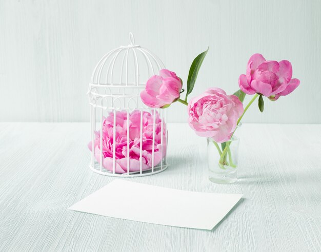 White bird cage twith petals on wooden table. Three peonies flowers in glass vase. Empty invitation card for marriage celebration.